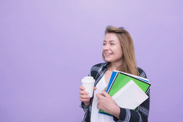 Smiling student girl stands on a purple background with notebooks and a cup of coffee in his hands and looks to the side. Happy girl with books and notebooks isolated on a purple background.Copyspace - Photo, Image