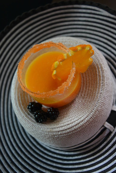 Ποτήρι με χυμό πορτοκάλι, καρότο ζάχαρη χείλος, ένα όμορφο κομμάτι του πορτοκαλί και τα βατόμουρα σε ένα άσπρο γυναικείο καπέλο με μαύρες ρίγες - Φωτογραφία, εικόνα