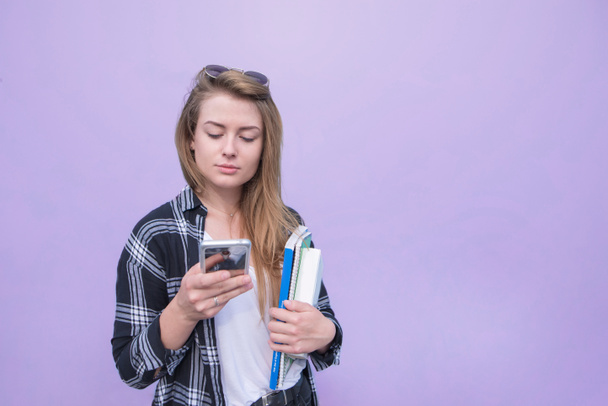 カジュアルな服を着ている深刻な女の子はスマート フォンを使用して、ノートブック、紫色の背景に本を保持します。女性は紫色の背景に分離されたスマート フォンを使用します。Copyspace - 写真・画像