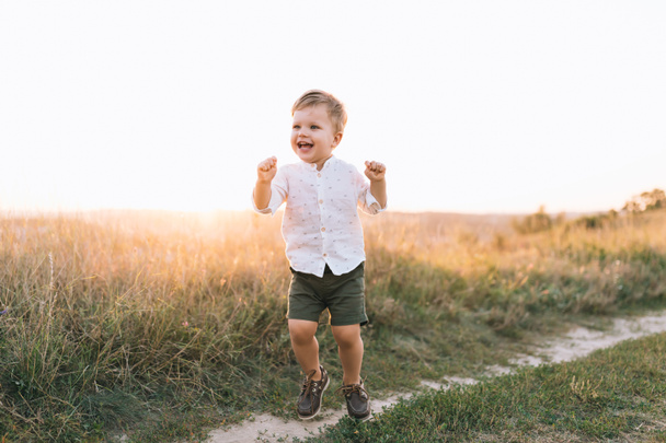 entzückend glücklicher kleiner Junge, der bei Sonnenuntergang auf einem ländlichen Pfad spaziert  - Foto, Bild