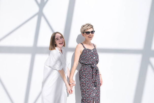 Two happy fashionable girls took each other by the hand and stand on the background of a white wall with shadows. Fashionable photo of two smiling girlfriends on the background of a white wall - Photo, Image