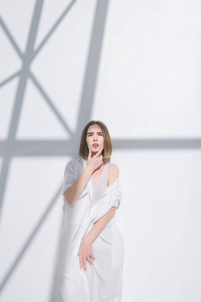 Стильная женщина-модель позирует на светлом фоне в белой одежде. Модная фотография стильной женщины на белом абстрактном фоне. Свет, тень, белая стена и стильная женщина
. - Фото, изображение