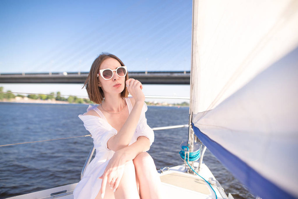 Portrait of an attractive woman sitting on a sailing yacht and looking at camera at the background of the river and bridge. Girl is resting on a yacht on a sunny, warm day - Фото, зображення