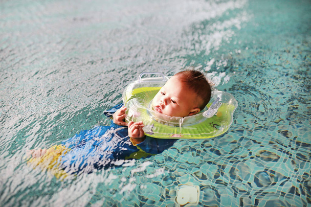 New born in swimming suit floating in pool with safety by baby neck floats. - Zdjęcie, obraz