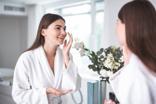 Portrait of positive lady applying cream on face while looking at reflection in cozy bathroom. She gesticulating hands. Treatment procedure concept - Photo, image