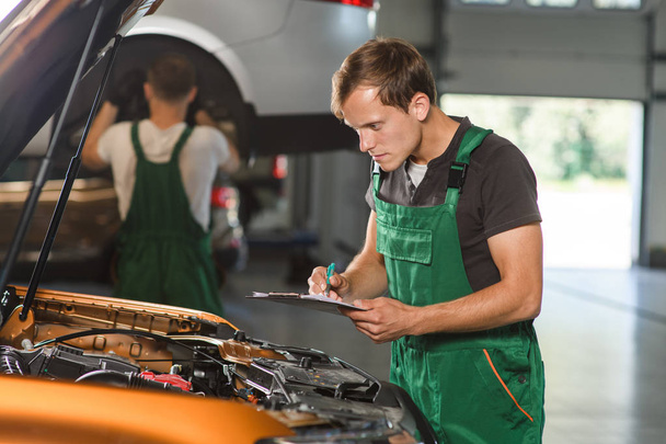 A young mechanic in green overalls is studying the cause of the car's malfunction at the service station - Photo, Image