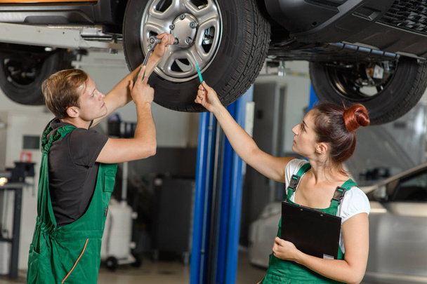 A beautiful and young girl locksmith conducts diagnostics of the suspension of the car with her colleague from the car-care center, they are dressed in green overalls - Photo, image