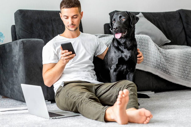 freelancer guy sitting at home working with a dog in an embrace, black labrador. - Fotoğraf, Görsel
