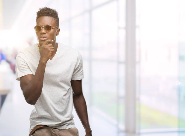 junger afrikanisch-amerikanischer Mann mit Sonnenbrille ernstes Gesicht über Frage nachdenkend, sehr verwirrte Idee - Foto, Bild