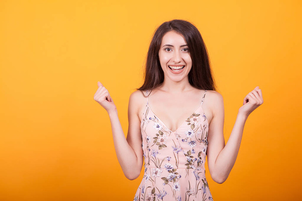 Attractive young woman smilling at the camera in studio over yellow background in short dress - Photo, Image