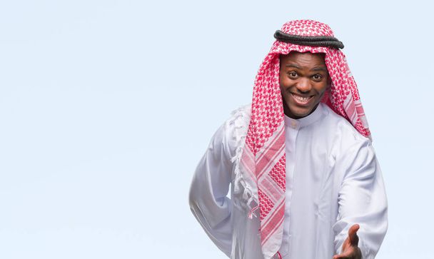 Молодой арабический африканский мужчина, одетый в традиционную кеффию на изолированном фоне, улыбаясь, дружелюбно предлагая рукопожатие в качестве приветствия и приветствия. Успешный бизнес
. - Фото, изображение