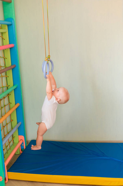 Lindo bebé colgando de anillos en el gimnasio de la pared del hogar. Concepto de desarrollo deportivo temprano del bebé
 - Foto, Imagen