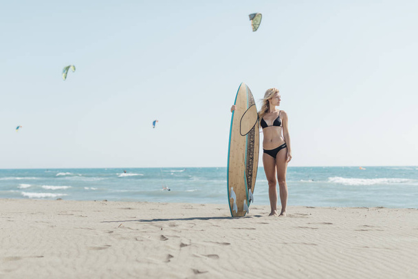 Portrait of beautiful young Caucasian girl standing at beach with her surf board. - Photo, image