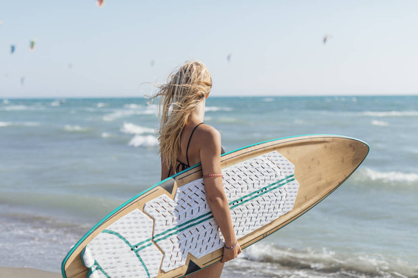 Back view of beautiful young Caucasian girl standing at beach with her surf board. - Photo, image