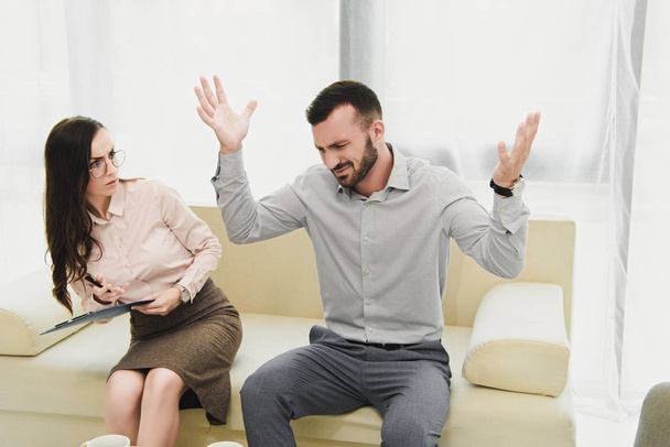 female psychiatrist with clipboard listening emotional patient gesturing in office - Fotó, kép