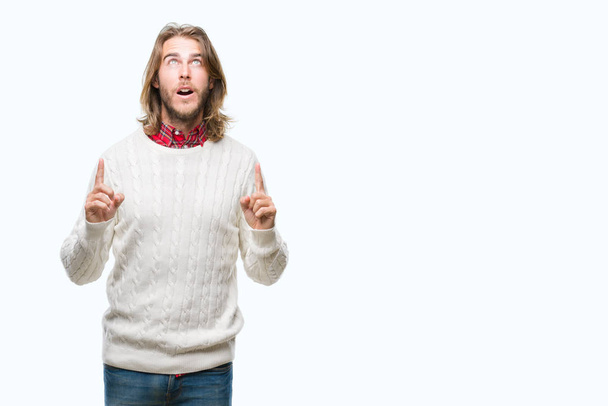 Young handsome man with long hair wearing winter sweater over isolated background amazed and surprised looking up and pointing with fingers and raised arms. - Photo, Image
