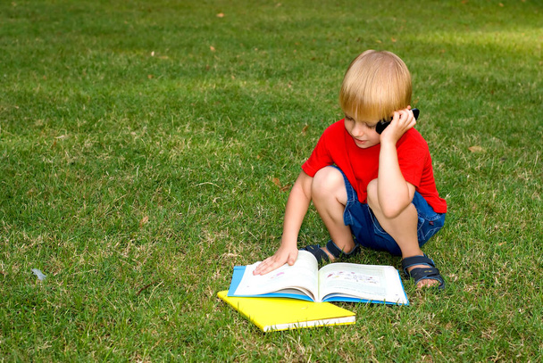 милый маленький мальчик сидит на зеленой траве с книгами и разговаривает по мобильному телефону
 - Фото, изображение