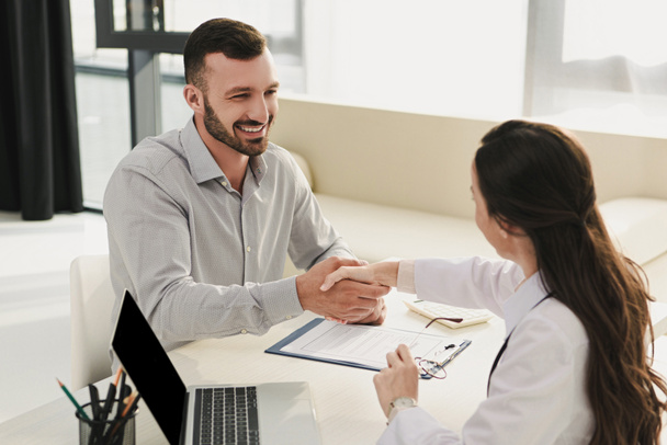 smiling client shaking hands with doctor in office with laptop and insurance claim form - Photo, Image