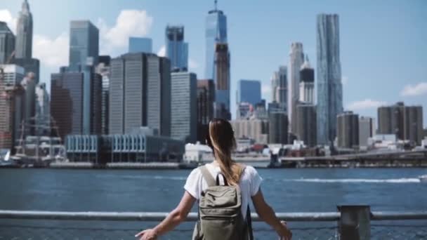 Back view of young happy tourist girl with backpack and arms wide open with joy at famous Manhattan skyline in New York - Imágenes, Vídeo
