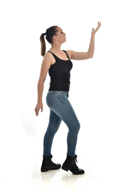 黒シングルとジーンズを身に着けているブルネットの女の子の完全な長さの肖像画。立ちポーズ、横顔。ホワイト スタジオの背景に分離. - 写真・画像