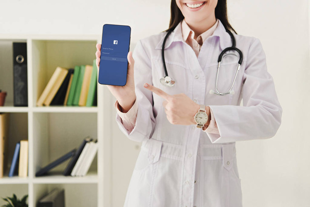 cropped view of doctor with stethoscope pointing at smartphone with facebook app - Φωτογραφία, εικόνα