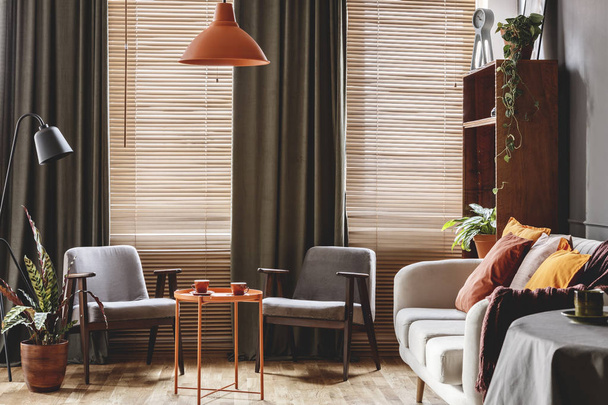 Grey armchair at orange table in dark retro living room interior with drapes and blinds - Fotoğraf, Görsel