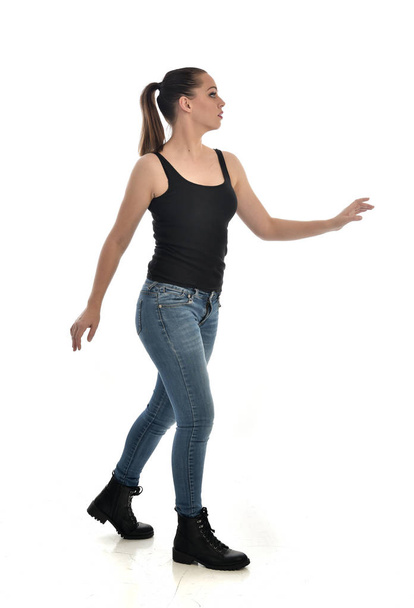 full length portrait of brunette girl wearing black single and jeans. standing pose, side profile. isolated on white studio background. - Foto, Bild