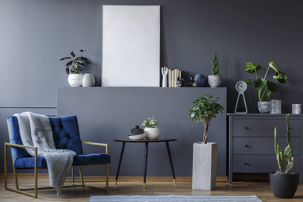 Real photo of a monochromatic living room interior with a blue armchair with a blanket standing next to a table and a shelf with an empty poster, with plants and ornaments around - Fotó, kép