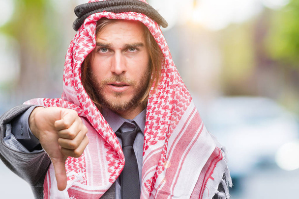 Young handsome arabian man with long hair wearing keffiyeh over isolated background looking unhappy and angry showing rejection and negative with thumbs down gesture. Bad expression. - Photo, Image