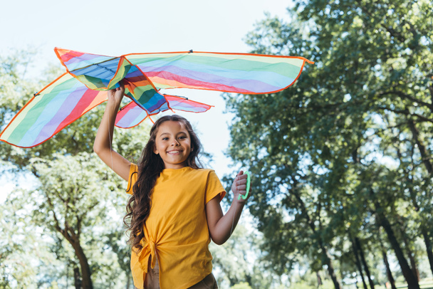 low angle view of happy child holding colorful kite and smiling at camera in park - Photo, Image