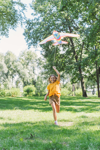 beautiful happy child holding colorful kite and smiling at camera while running on grass in park - Fotó, kép