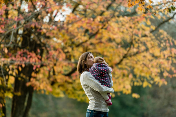 Jeune femme se tenait avec bébé fille dans les bras sous l'arbre d'automne embrassant sa tête
. - Photo, image