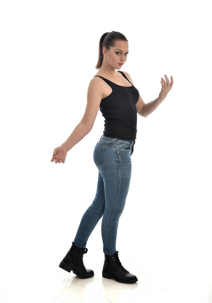 full length portrait of brunette girl wearing black single and jeans. standing pose, side profile. isolated on white studio background. - 写真・画像