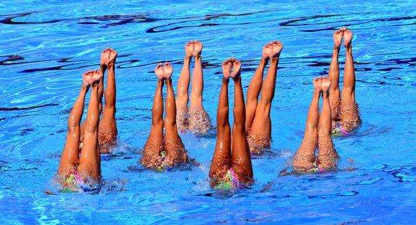 Синхронные пловцы указывают вверх из воды в действии. Синхронное движение ног пловцов. Синхронное плавание команды, выполняющей синхронную рутину сложных движений в воде
. - Фото, изображение