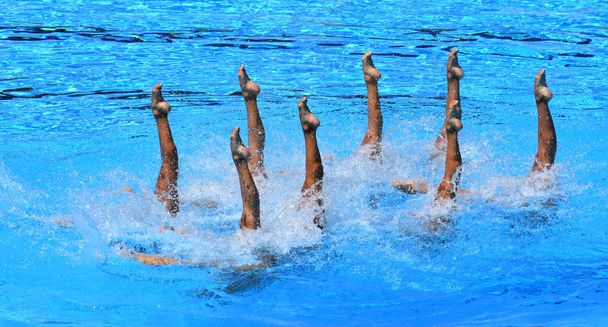 Синхронные пловцы указывают вверх из воды в действии. Синхронное движение ног пловцов. Синхронное плавание команды, выполняющей синхронную рутину сложных движений в воде
. - Фото, изображение