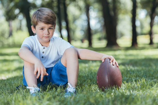 aranyos kisfiú gazdaság rögbi labdát, miközben ül a fűben, és néztem a kamera - Fotó, kép