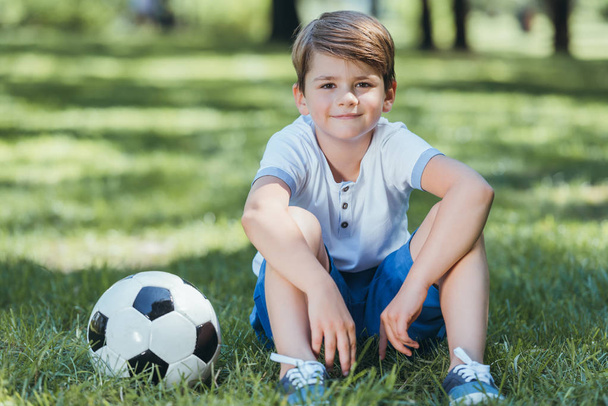 lindo niño sentado en la hierba con pelota de fútbol y sonriendo a la cámara en el parque
  - Foto, Imagen