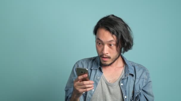 Young bearded male hipster looking at his phone to find great new from mobile message application. Bored Asian man looking at mobile phone and extremely happy as he see good news with blue background. - Video