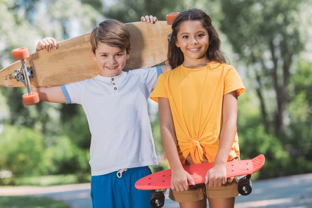 cute happy kids holding skateboards and smiling at camera in park - Φωτογραφία, εικόνα