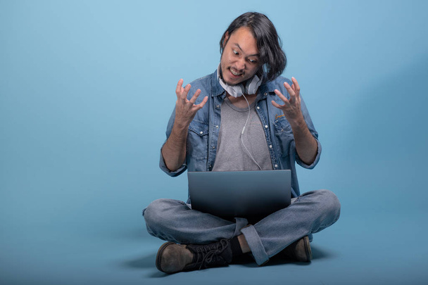 El joven barbudo se sienta en el suelo usando una computadora en un fondo azul. Cansado joven hipster asiático utilizando el ordenador portátil, tiro de cuerpo completo. Concepto de trabajo hipster generación joven
. - Foto, Imagen