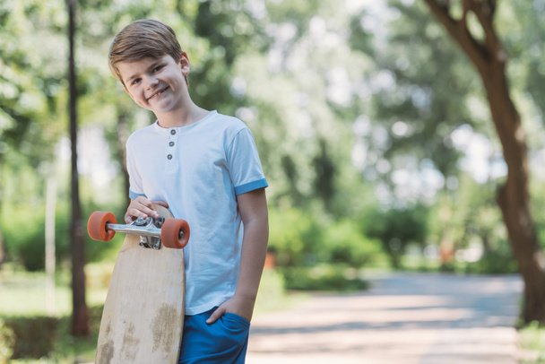 happy kid standing with skateboard and smiling at camera in park   - 写真・画像
