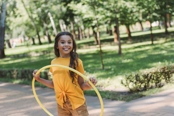 lindo niño feliz sosteniendo hula hoop y sonriendo a la cámara en el parque
  - Foto, imagen