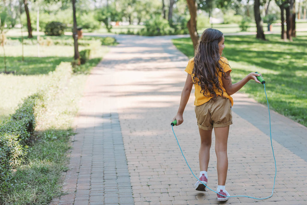 rear view of cute kid holding jumping rope and walking in park  - Fotó, kép