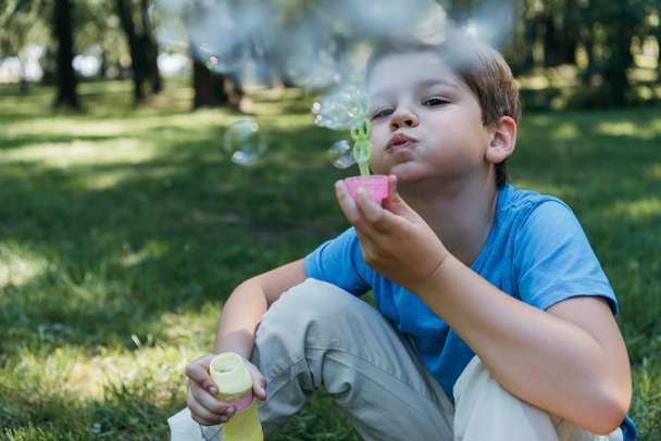 adorable child blowing soap bubbles while sitting on grass in park - Photo, Image