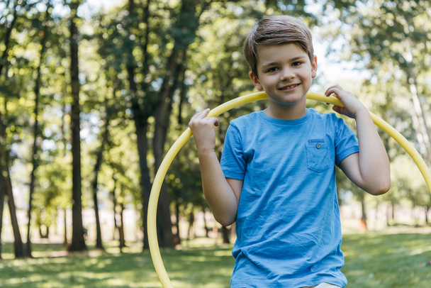 adorable happy boy holding hula hoop and smiling at camera in park - 写真・画像