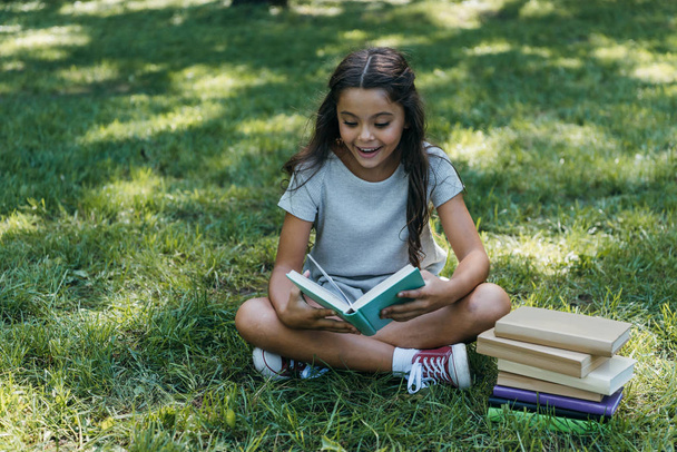 adorable happy child sitting on grass and reading book in park - Fotografie, Obrázek