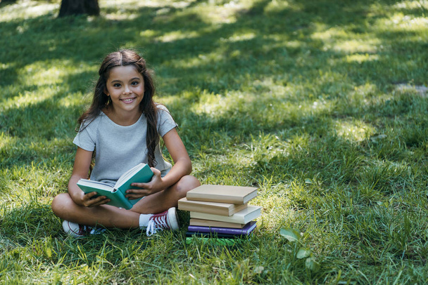 cute happy child sitting in grass with books and smiling at camera in park - Фото, изображение