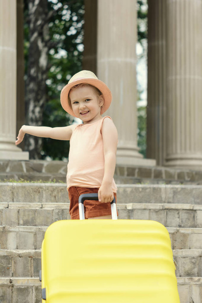 Een klein meisje met blond haar staat op de trappen met een grote gele koffer. Een mooi kind in een stro hoed hief een hand. - Foto, afbeelding