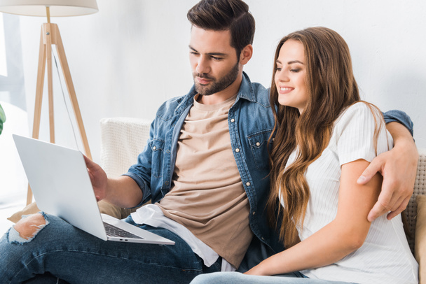 handsome man pointing by finger on laptop screen to girlfriend on sofa at home - Φωτογραφία, εικόνα