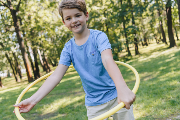 happy boy playing with hula hoop and smiling at camera in park - Photo, Image